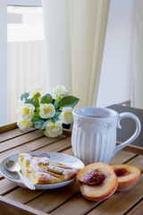 piece of peach pie on a white plate, white mug with tea on a wooden surface and yellow flowers in the background, natural light from the window. Vertical photo