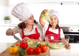 young mother and little daughter at house kitchen preparing salad for lunch wearing apron and cook hat