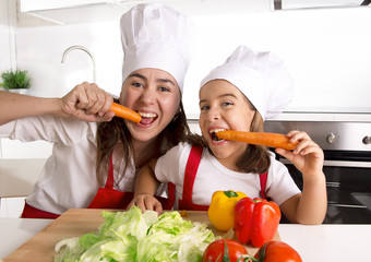 happy mother and little daughter in apron and cook hat eating carrots together 