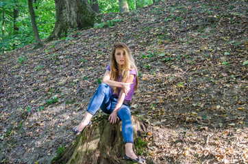 Young beautiful woman model with long hair in jeans and a tank top walks through the forest park among trees and vegetation posing on a stump of felled trees