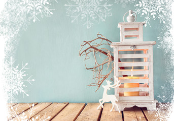 white wooden vintage lantern with burning candle, wooden deer, christmas gifts and tree branches on wooden table. retro filtered image with glitter and snowflake overlay
