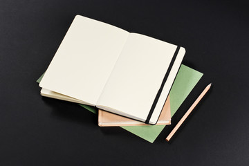 Open blank notepad with empty white pages laying above book with pencil on black background. ideal for text