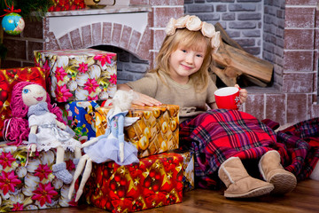 The little girl  is sitting at a fireplace and Christmas gifts. The girl covered by a plaid  is holding a red mug in her hand.