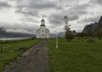 Fototapeta na wymiar A walk way to the front of the church with some gravestones and light poles. Mountains and low haning clouds