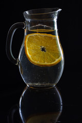 Jug with water and one slice of orange on black background