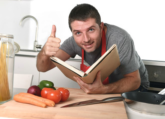 young happy man at kitchen reading recipe book in apron learning cooking