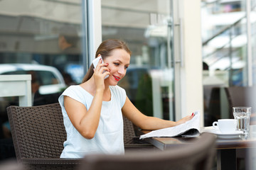 Young woman reading a newspaper and talking on a cell phone sitt