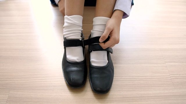 Asian Thai schoolgirl student in high school uniform is wearing her black shoes in cute education fashion design on the wooden floor classroom 