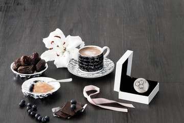Silver ring in gift box with pink ribbon in morning coffee setting with dry dates, orange marmelade, chocolate, blueberries and white lily flower