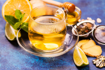 Hot green tea in a glass cup served with lemon, ginger, lime, honey and mint on grunge blue wooden background. Selective focus