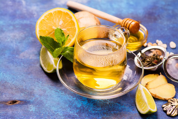 Hot green tea in a glass cup served with lemon, ginger, lime, honey and mint on grunge blue wooden...