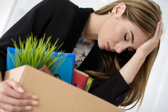 Young dismissed female worker in office holding carton box with