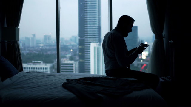 Young businessman watching movie on tablet computer sitting on bed at home
