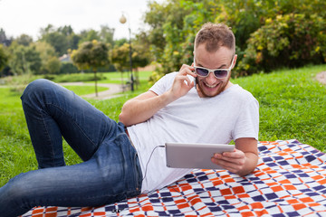Fototapeta premium Bearded young man lying in park on blanket. He is using white tablet pc.