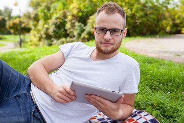 Bearded young man lying in park on blanket. He is using white tablet pc.