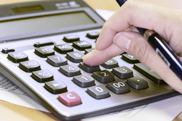Photo of hand holding pen and pressing calculator buttons over documents