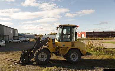 A small tractor parked with a blue sky with some clouds in the background, some cars in front of a building