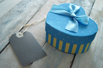 Heart shaped gift box decorated with a blue ribbon and a label, all on a white wooden table. Empty copy space for editor's text.