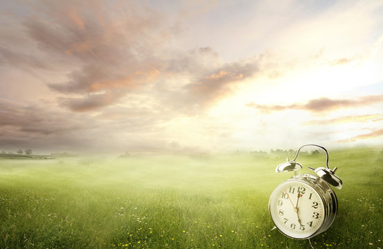 Alarm clock in green field and sunny sunrise sky. Daylight saving time. Springtime