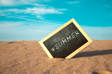 closeup of a chalkboard with a wooden frame and the word #summer written in it, placed on the sand of a beach