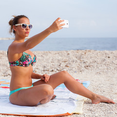 Pretty girl sitting on the beach and taking selfie by her phone.