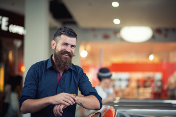 Stylish bearded man at the mall looking at the clock and laughing on the background of shop windows