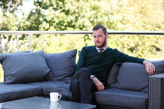 A handsome man in a shirt, tie and sweater sitting down outside on a summer day holding a mug. Relaxed and laid back.