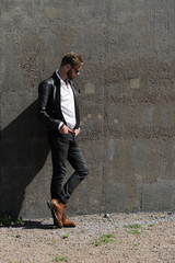 An attractive man wearing a black leather jacket and sunglasses leaning against a stone wall...