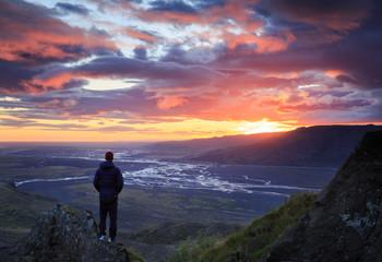Man standing on a ledge of a mountain, enjoying the beautiful sunset over a wide river valley in Thorsmork, Iceland.

