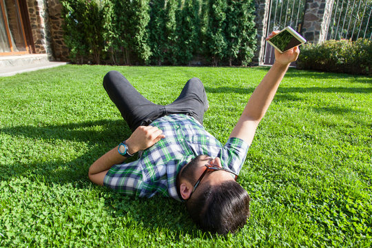 One young fashion middle eastern man with beard and fashion hair style is lying on a grass in a park taking selfie. holding smart phone, tablet, camera and  looking at camera.