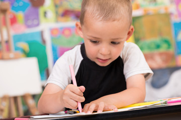 Cute and serious toddler boy is at the Art class. Looking down and painting with gray felt pen. Colorful wall and easel in background.