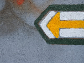 Yellow tourist arrow pointing left on a grey concrete wall (the wall is painted by red graffiti signs).