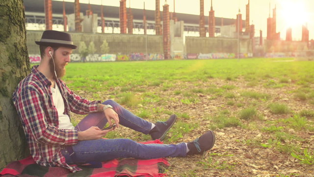 Young hipster guy relaxing listening music laying down next to a tree in a park.