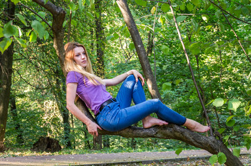 Young beautiful sexy girl model of European appearance with long hair in a shirt and jeans sitting on a tree during a walk in the autumn sunny park forest of trees and vegetation