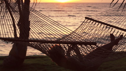 Young Woman with Blonde Hair Using Mobile Smart Phone while Relaxing in a Hammock by the Ocean Between Palm Trees at Sunset in Hawaii. Beautiful Sunset Silhouette Warm Instagram Color Tone