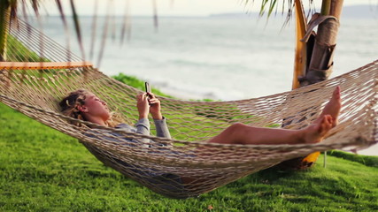 Revealing Pan of Beautiful Blonde Young Woman with Curly Hair Using Mobile Smart Phone while Relaxing in a Hammock by the Ocean Between Palm Trees at Sunset in Hawaii. 