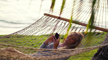 Young Woman Laughing Using Mobile Smart Phone while Relaxing in a Hammock by the Ocean Between Palm Trees at Sunset in Hawaii.  Instagram Color Tone