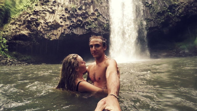 Cute Young Couple Holding Kissing in front of Waterfall with POV Selfie Stick. Happy cheerful young multicultural couple on travel. Lush Green Waterfall in Hawaii.