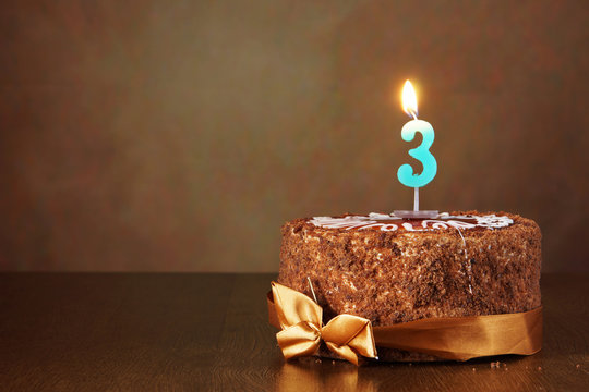 Birthday chocolate cake with burning candle as a number three on brown background