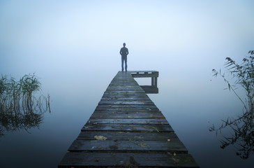 Fototapeta premium Mężczyzna stojący na molo nad jeziorem w mglisty, szary poranek.