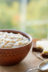 Oatmeal porridge in bowl on wooden table near window, rustic breakfast. Bread and cheese on side