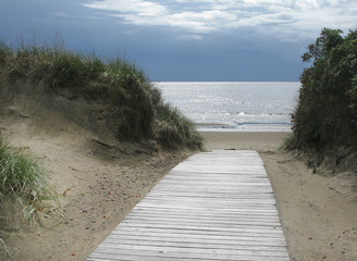 Sand dune landscape with wooden boardwalk leading towards sea and sky at Skrea Strand on a sunny day with dark clouds in Falkenberg, Sweden.