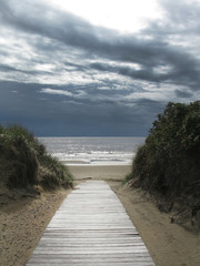 Sand dune landscape with wooden boardwalk leading towards sea and sky at Skrea Strand on a sunny day with dark clouds in Falkenberg, Sweden.