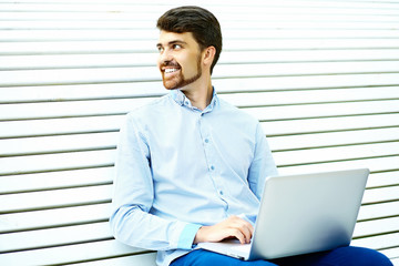 Young handsome smiling businessman model sitting on the park bench using laptop in casual hipster cloth