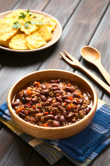 Wooden bowl of chili con carne with homemade tortilla chips in the back, photographed with natural light (Selective Focus, Focus in the middle of the chili)