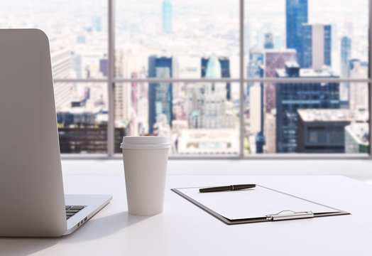 A workplace in a modern panoramic office in Manhattan, New York City. A laptop, notepad and a coffee cup are on the white table. 3D rendering. Toned image.