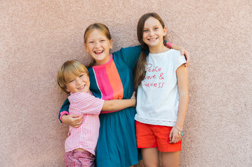 Outdoor portrait of 3 cute kids, wearing colorfil clothes, standing aginst pink wall