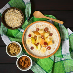 Oatmeal porridge with grape, apple and banana in wooden bowl, walnut, almond and raw oatmeal on the side, photographed overhead on dark wood with natural light