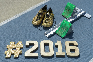 Large gold hashtag 2016 message with matching shoes next to starting blocks on a textured blue and...
