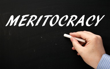 Hand writing the word Meritocracy in white text on a blackboard. A situation where people achieve...